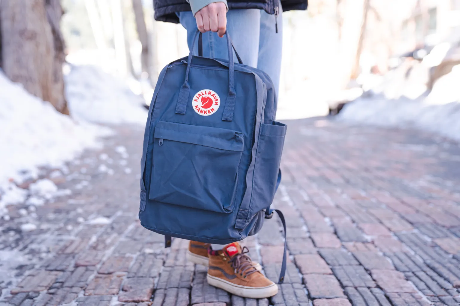 在积雪的人行道上,人们手提着 Fjallraven Kanken 背包的顶部提手。 在积雪的人行道上,人们手提着 Fjallraven Kanken 背包的顶部提手。